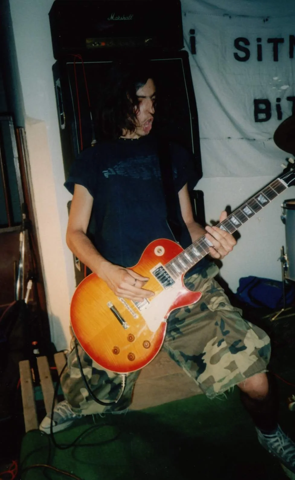 A young man with long dark hair, wearing a black T-shirt and camouflage shorts, playing an orange electric guitar with a burst finish. He is standing on a wooden floor, with Marshall speakers behind him and a white banner with partially visible text 