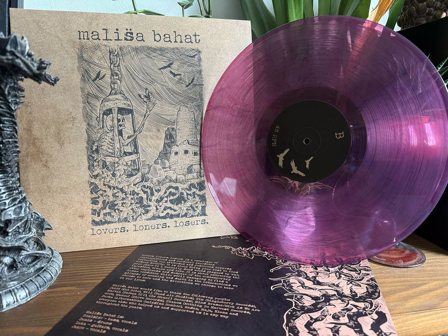 A purple vinyl record on display next to a beige album cover with black and white artwork and text, and a black sheet of paper with text on a wooden surface.