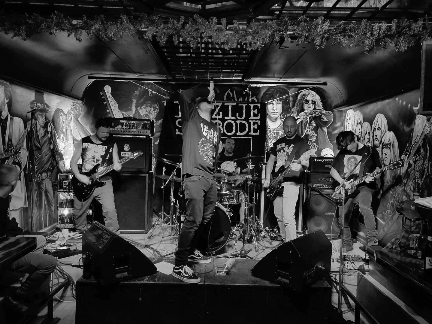 A black and white photo of a band performing live on stage in a small venue. There are five band members—one singing into a microphone, two playing guitars, one on drums, and one on bass guitar. 