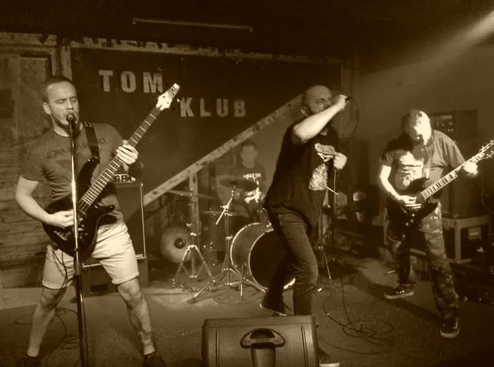 A band performing live on stage at a club with a sign in the background reading "TOM KLUB." Four musicians are visible: two guitarists, a vocalist, and a drummer.