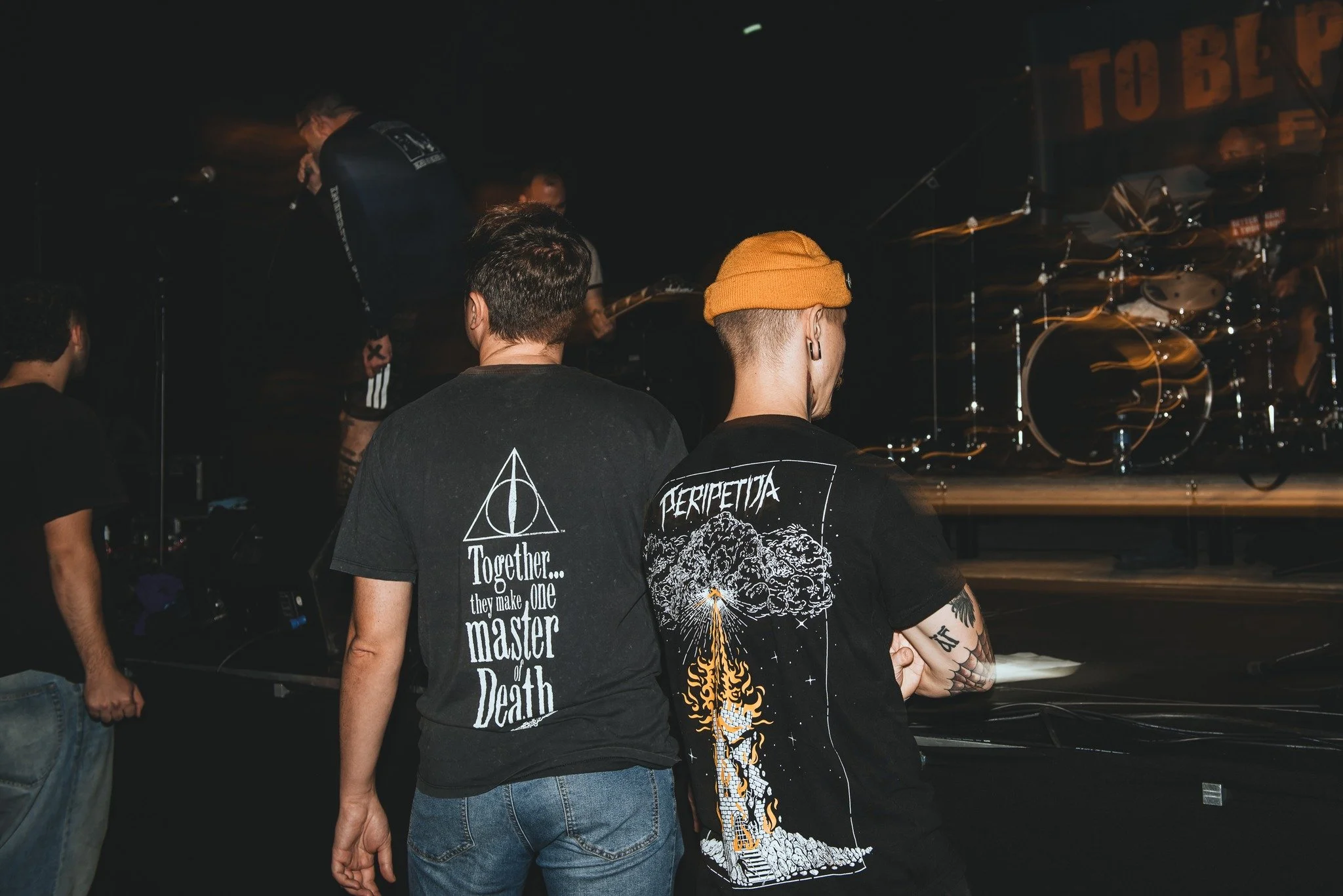 Three people standing and watching a stage with a drum set, at a concert or music event.