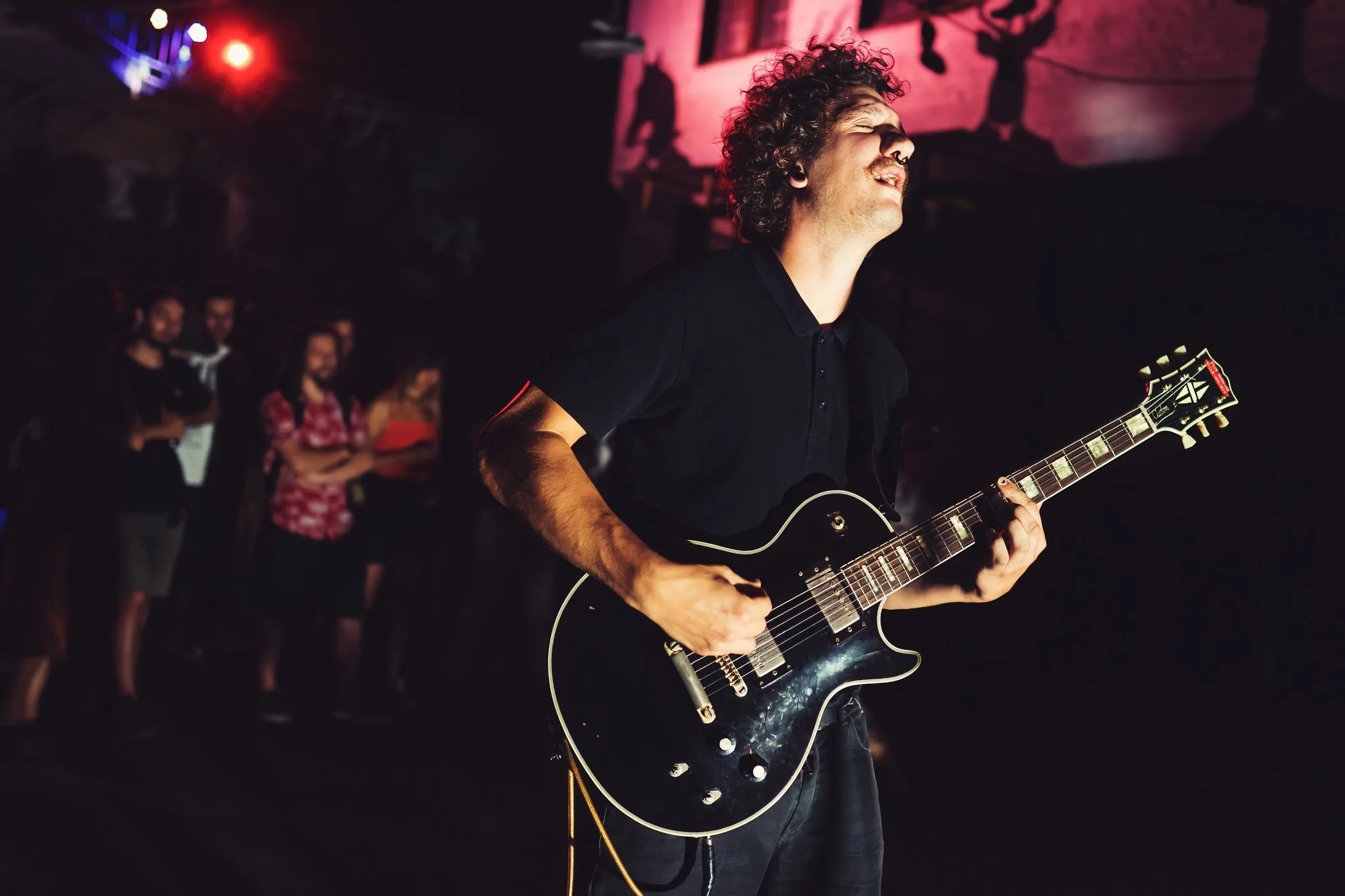 A musician with curly hair and a nose piercing playing a black electric guitar during a concert, with an audience watching in the background.