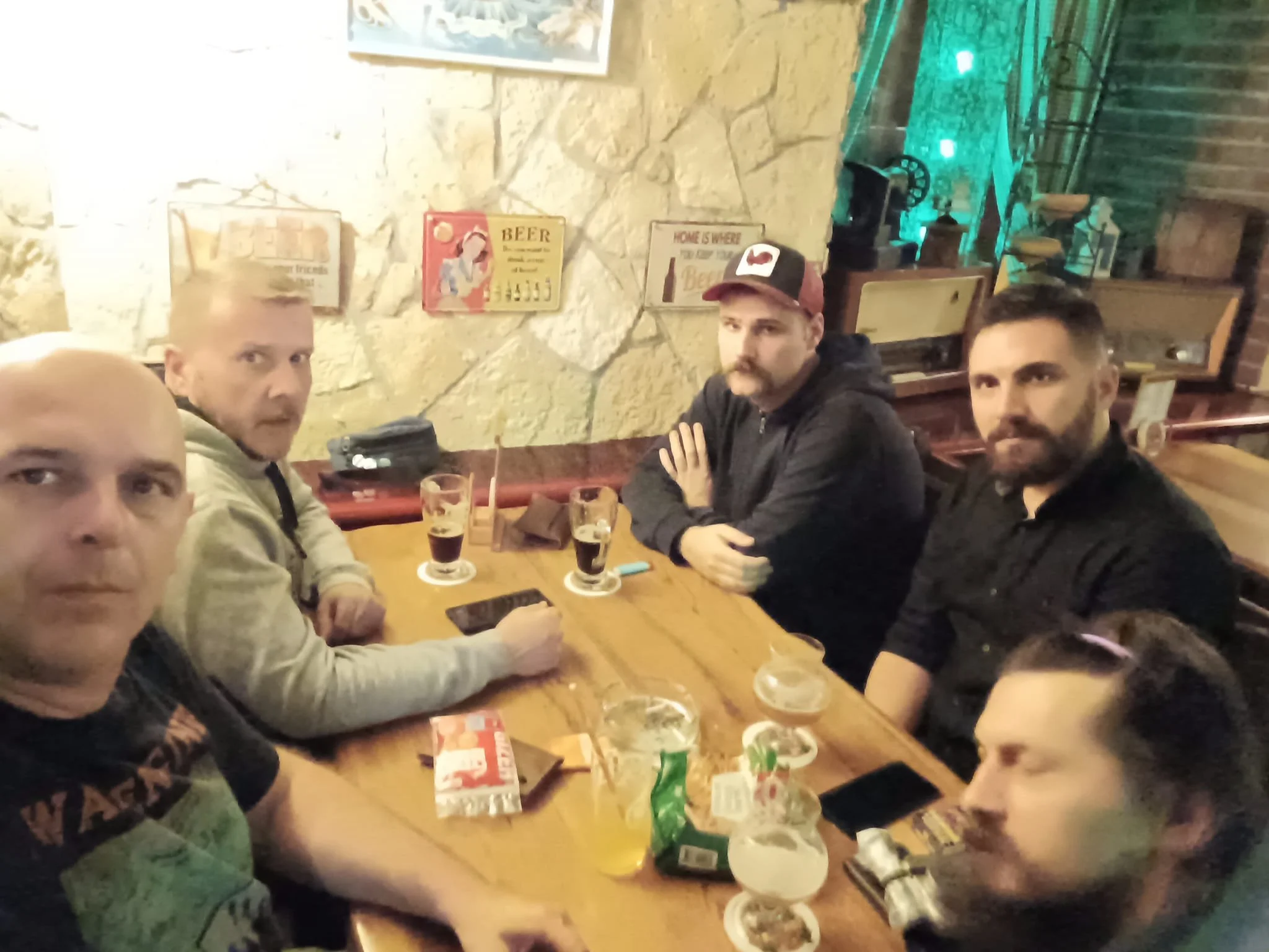 Four men sitting at a wooden table in a dimly lit restaurant or bar, with beers and snacks on the table, and a stone wall with framed pictures on it in the background.