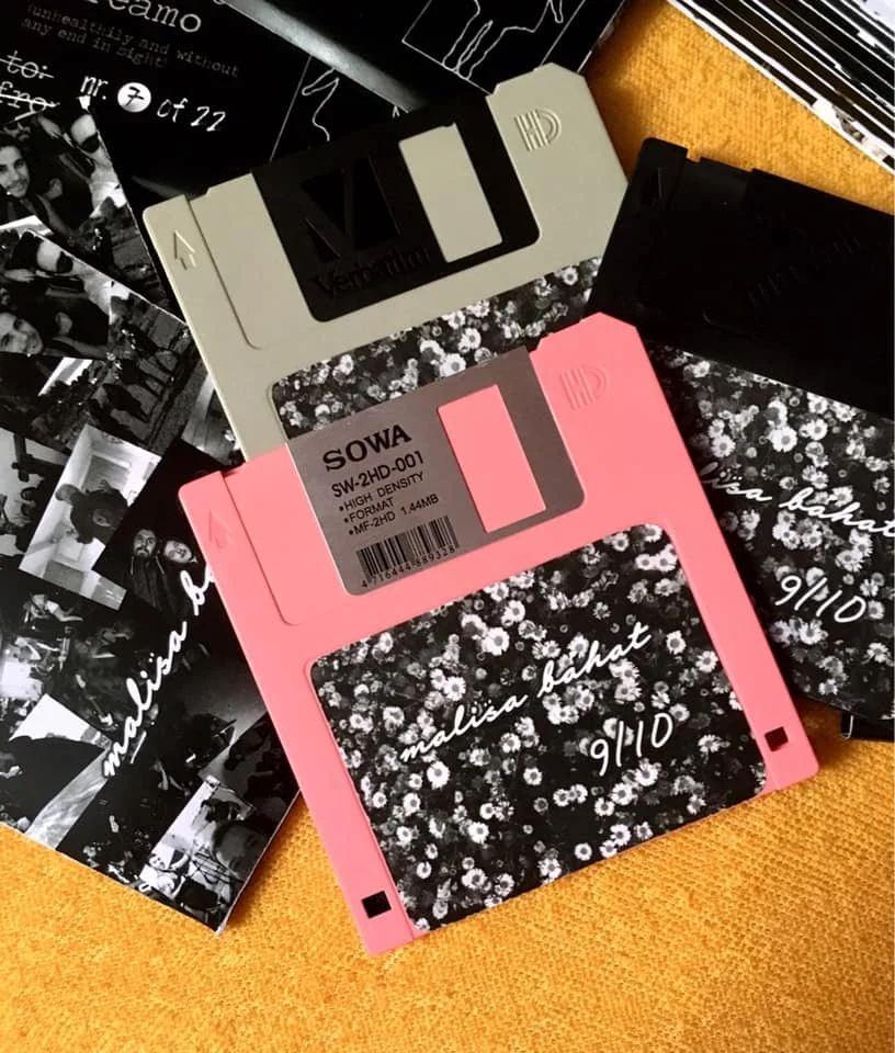 Two floppy disks, one pink labeled "MALISA BAHAT" and one beige labeled "MIX," are placed on top of black-and-white photographs and papers on an orange surface.