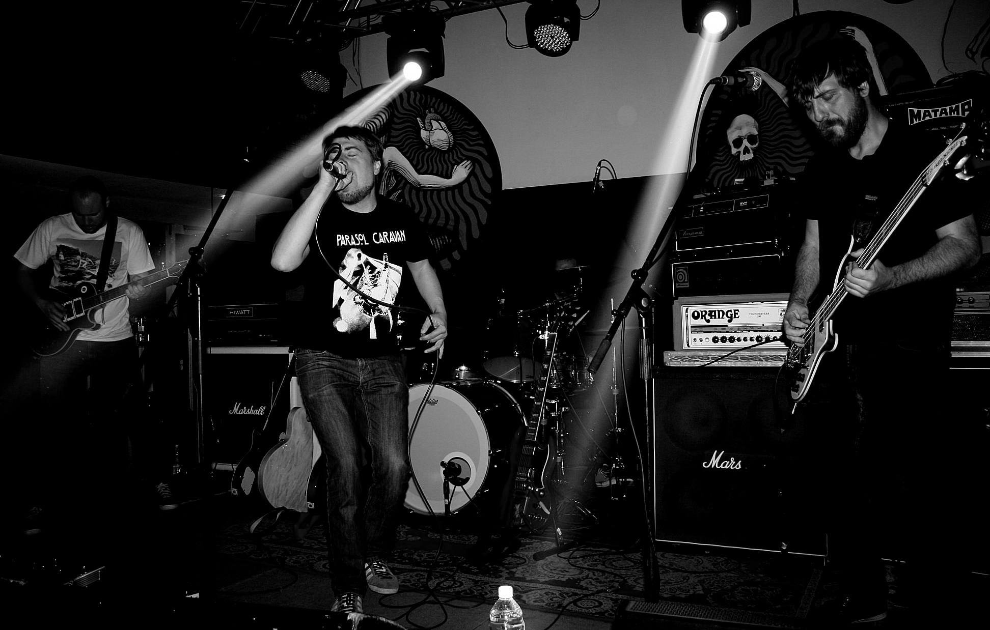 A black-and-white photo of a band performing on stage with three members visible. The lead singer, at the center, is singing into a microphone and holding a guitar. To the right, a guitarist is playing an electric guitar, and to the left, another mus