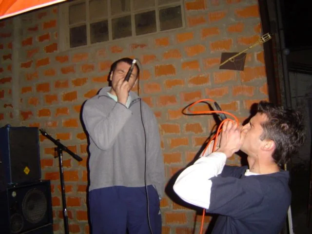 Two young men singing into microphones outdoors against a brick wall, with a speaker and microphone stand nearby.