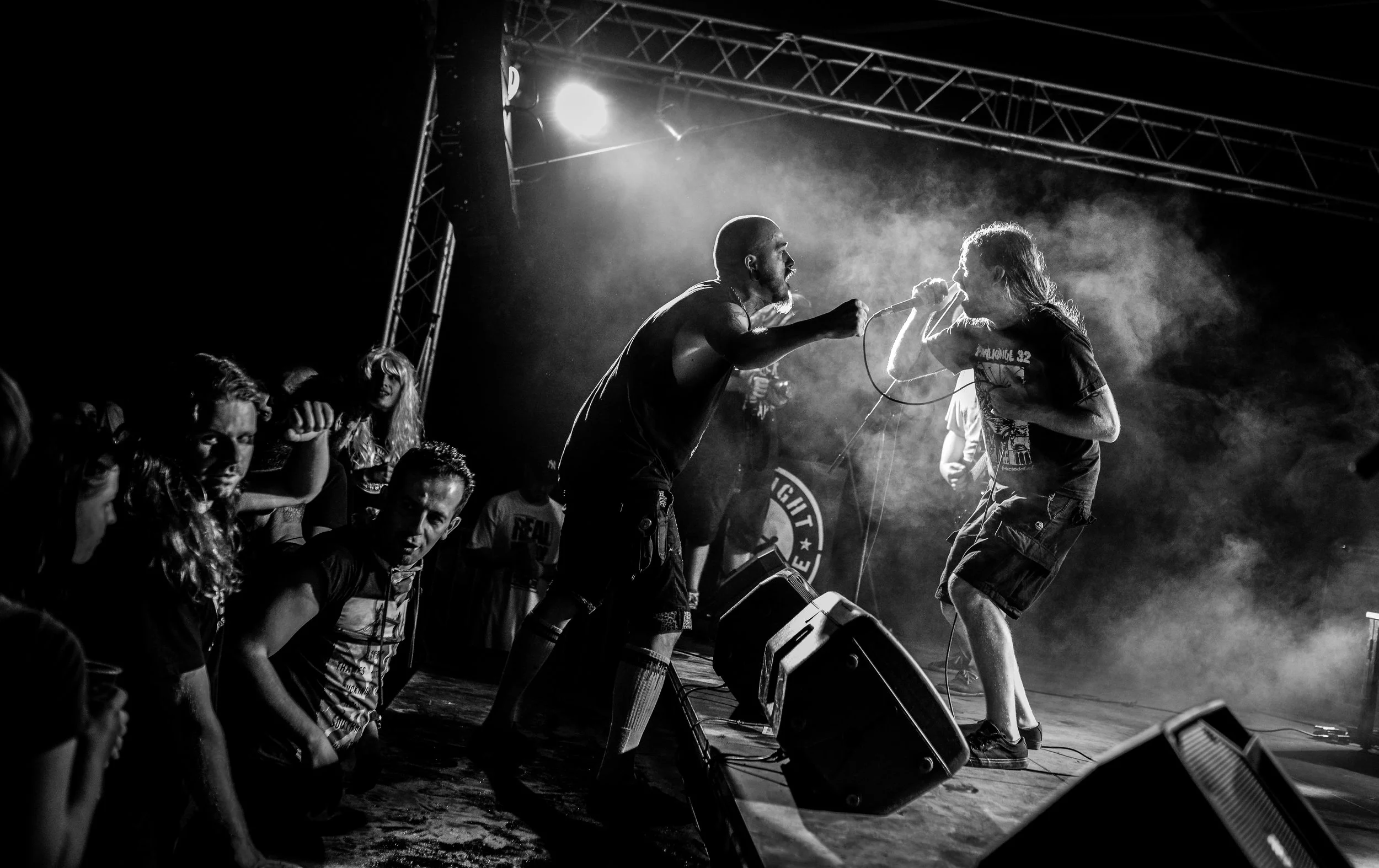 Black and white photo of a live music concert showing two male performers on stage, one singing into a microphone and the other holding the microphone, with an audience in front and stage lights shining down.