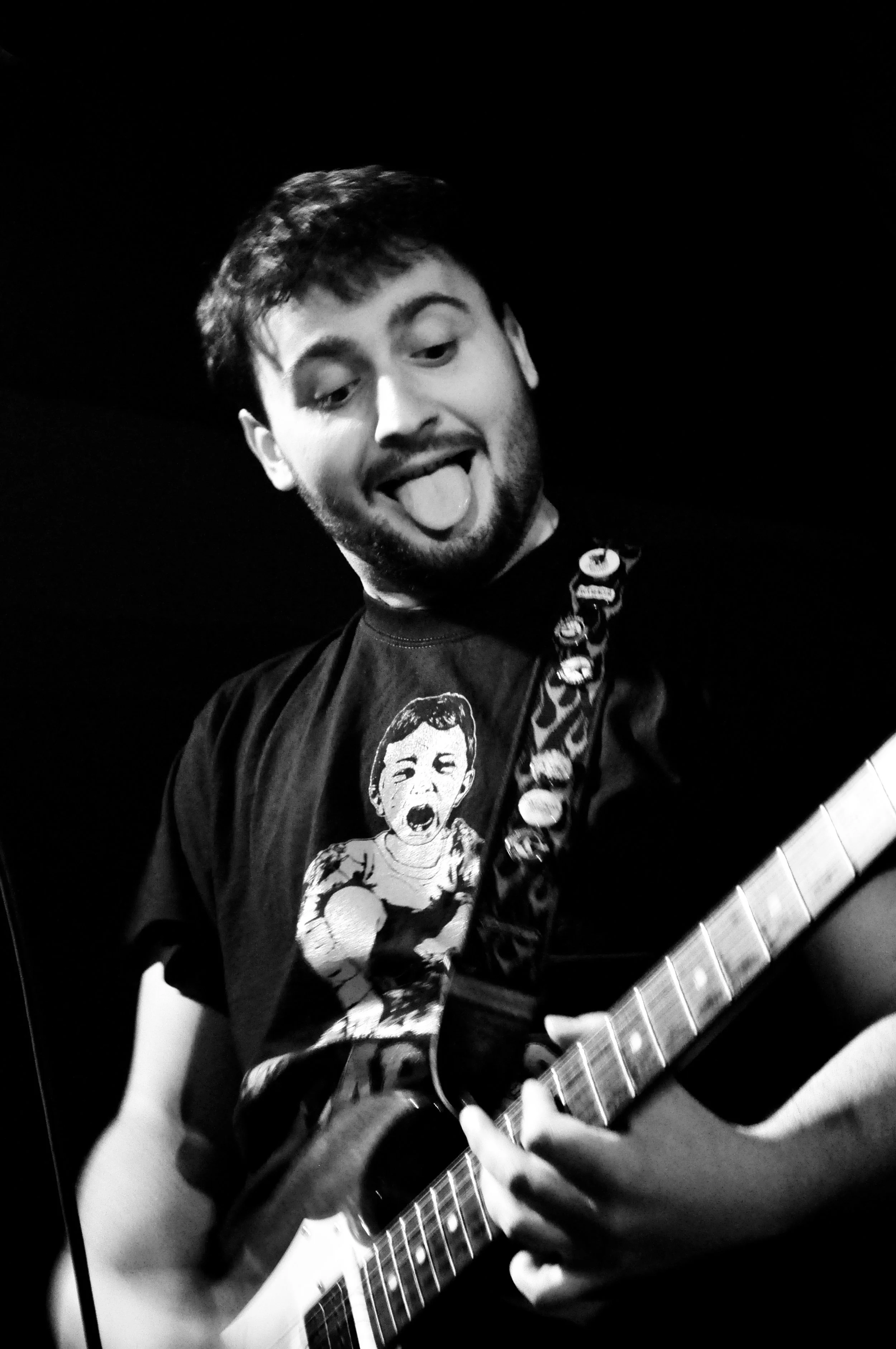 Black and white photo of a man with short hair and a beard, sticking out his tongue, playing an electric guitar, wearing a t-shirt with a cartoon graphic of a screaming boy.