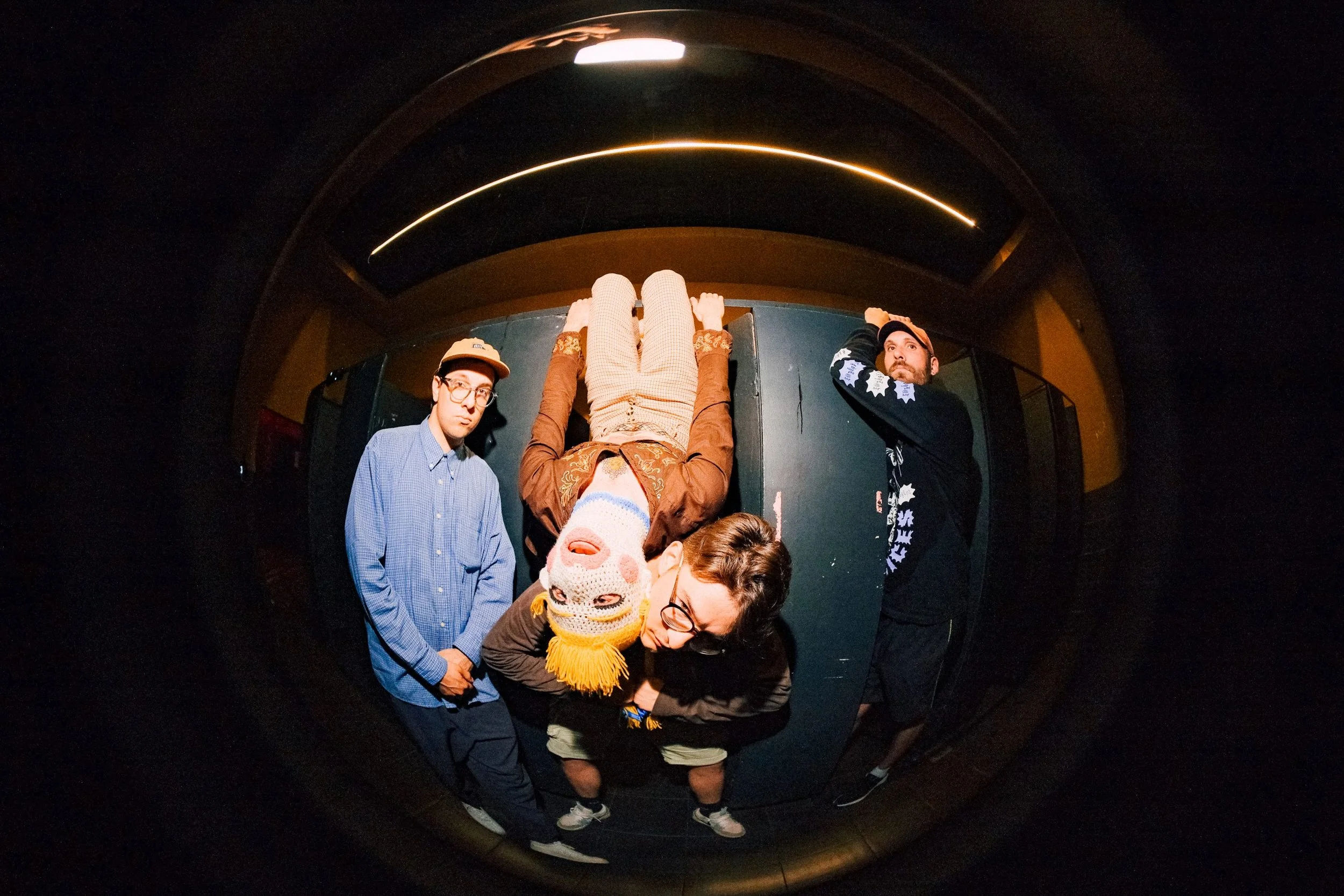 People standing inside a dark room, all four band members of SUPLEXX, viewed through a fisheye lens.