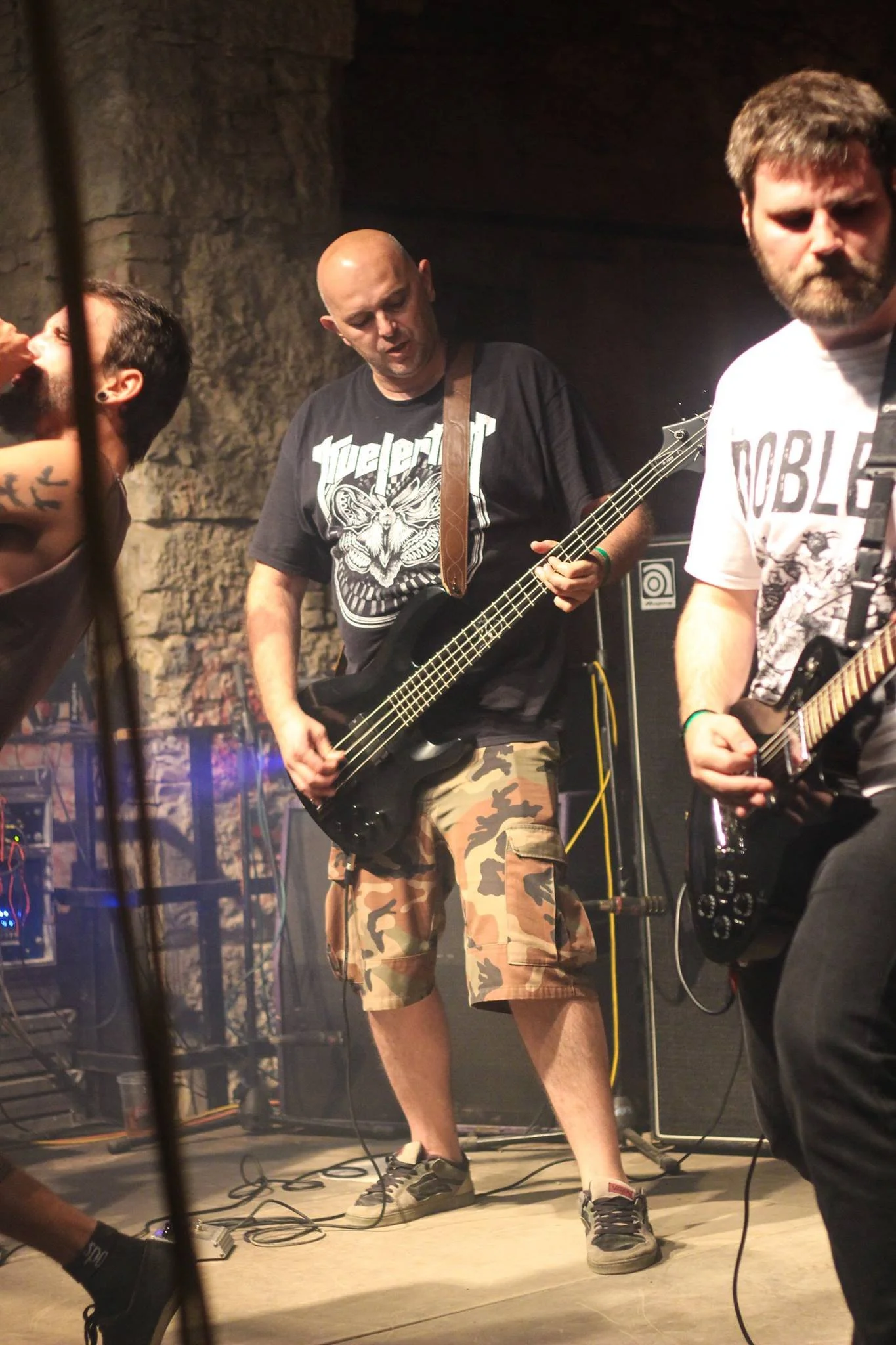 Four men playing musical instruments on stage, with a brick wall background and music equipment around.
