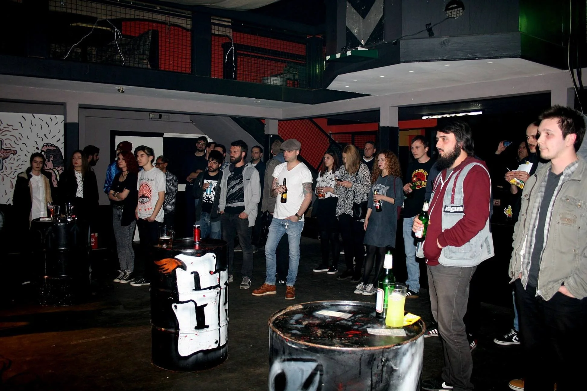 A group of people attending a social gathering or party in an industrial-style venue with black and red decor. Several individuals are holding drinks, and there are drums and bottles on tables in the foreground.