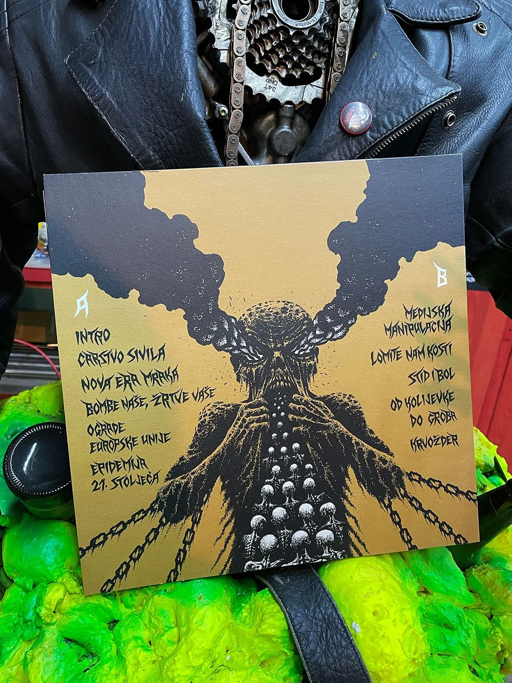 A black and yellow illustration of a skull with smoke or clouds billowing from the top, attached to a structure made of chains and skulls. The artwork is on a sign placed over a bright green object and is held by a person wearing a black leather jack