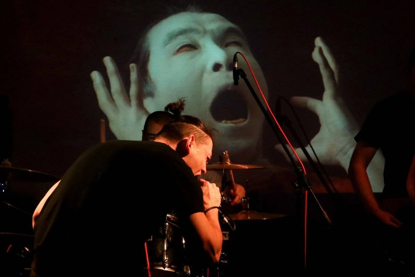 Musicians perform live on stage with a projection in the background showing a person's face with hands near the head and an open mouth.