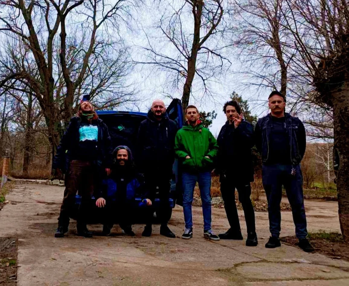 Group of six men standing outdoors on a cloudy day, with leafless trees in the background and a car behind them.