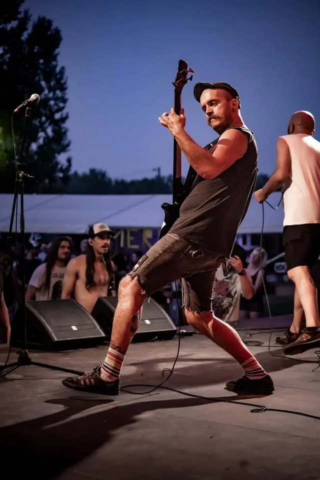 A musician playing an electric guitar on stage at an outdoor concert during dusk, with audience members in the background.