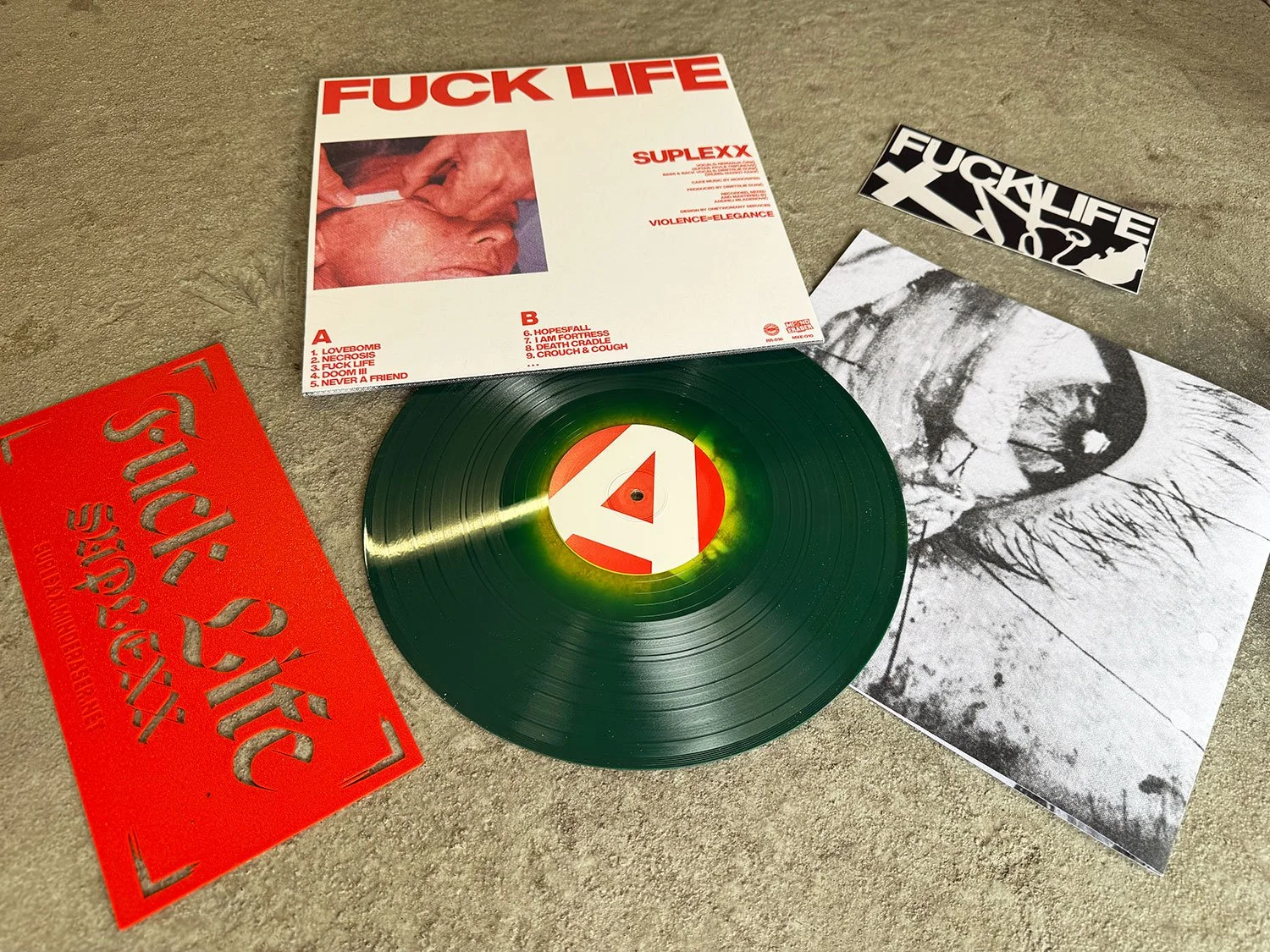A black vinyl record with a red, white, and yellow label featuring the letter A, placed on a gray surface. Surrounding the record are various album covers and posters, including a white cover with red text that says 'FUCK LIFE' and a tracklist, a bla