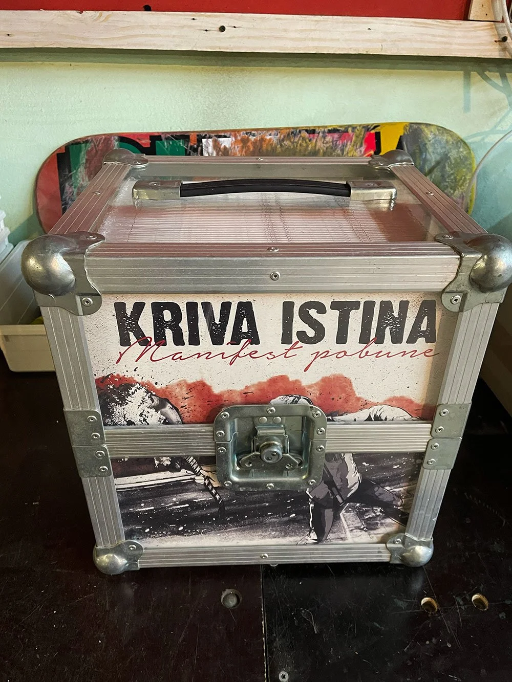 A metal box with a handle and latch, decorated with an image of a person holding a ski pole, and the words 'KRIVA ISTINA' and 'Manifest pobune' printed on it.