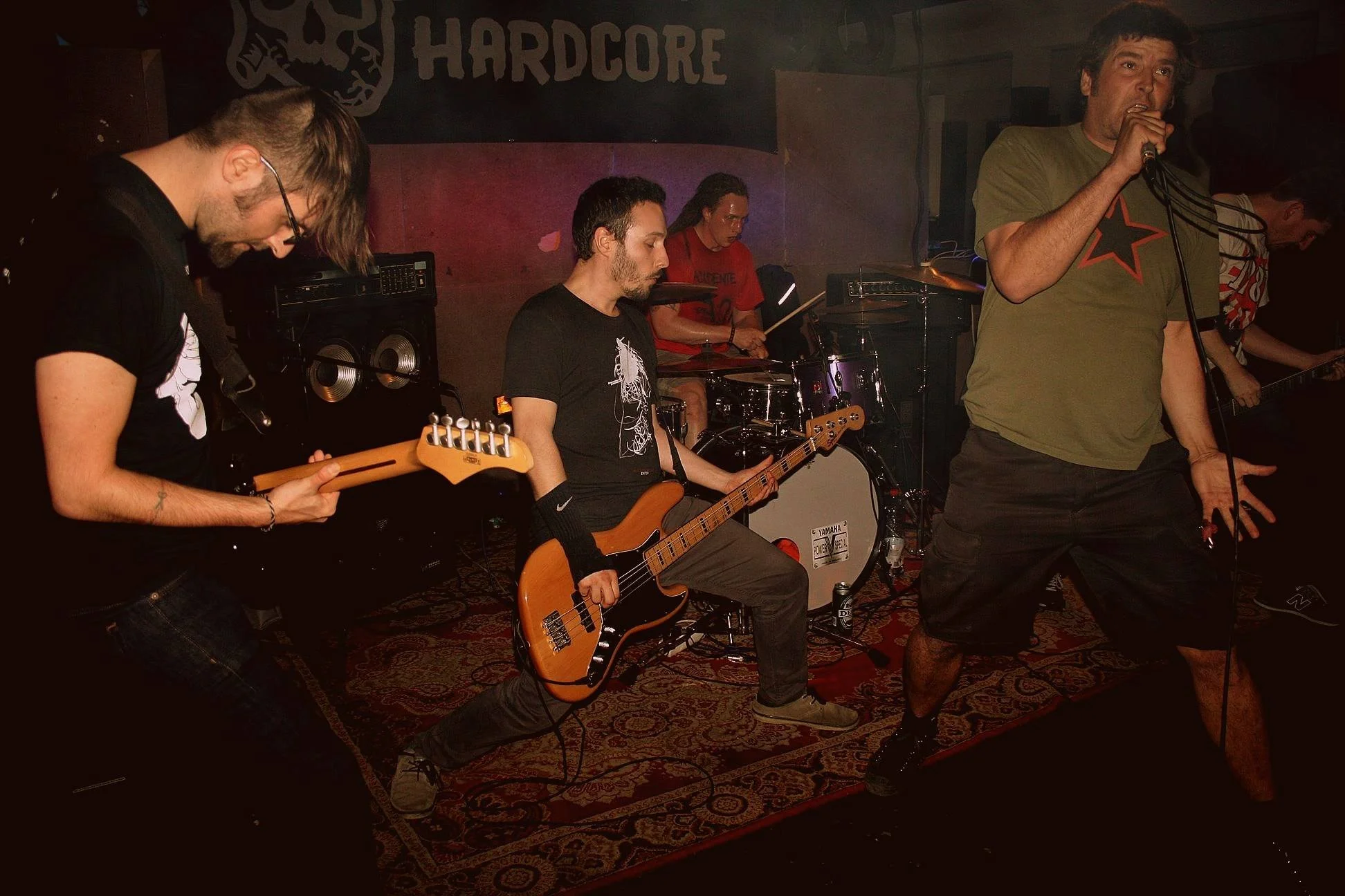 A band performs on stage in a dimly lit venue. The lead singer, wearing a green shirt with a star design, is singing into a microphone. To his left, a man plays the bass guitar, and another musician plays the electric guitar. A drummer in the backgro