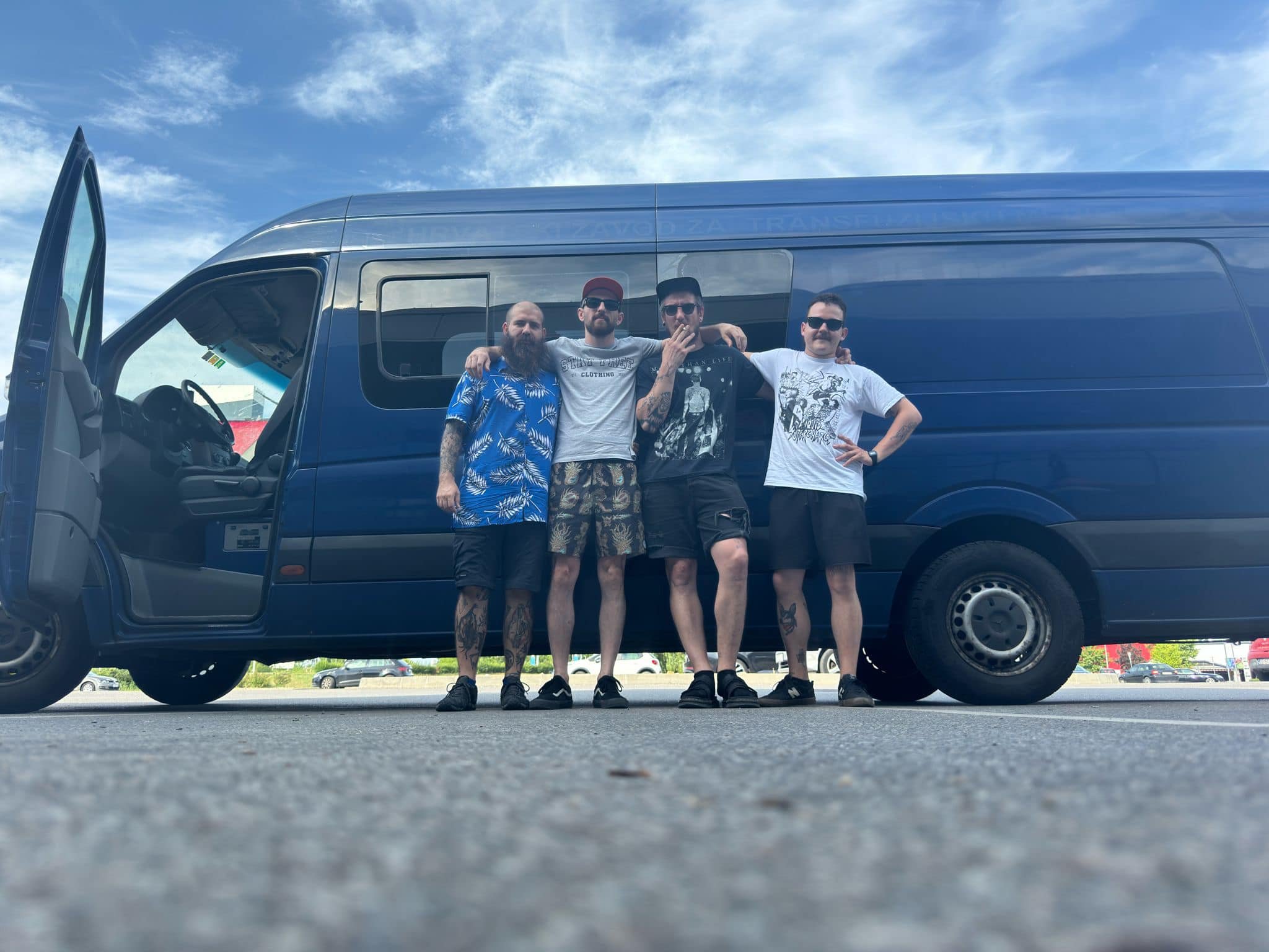 Four men stand arm in arm in front of a large blue van, with the photo taken from a low angle at ground level.