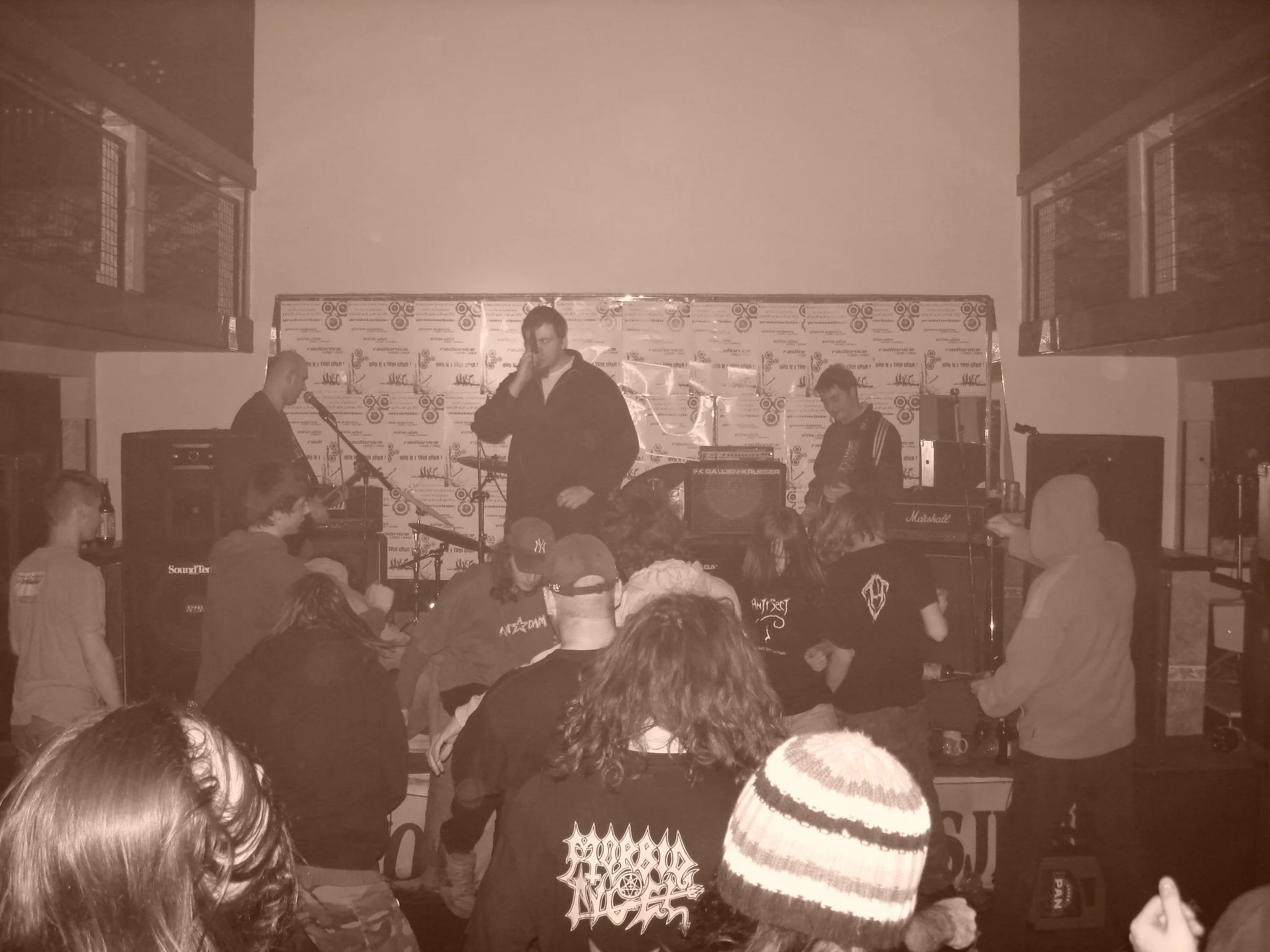 A band performing on stage with several musicians playing instruments and an audience watching. The background has promotional posters for the event.