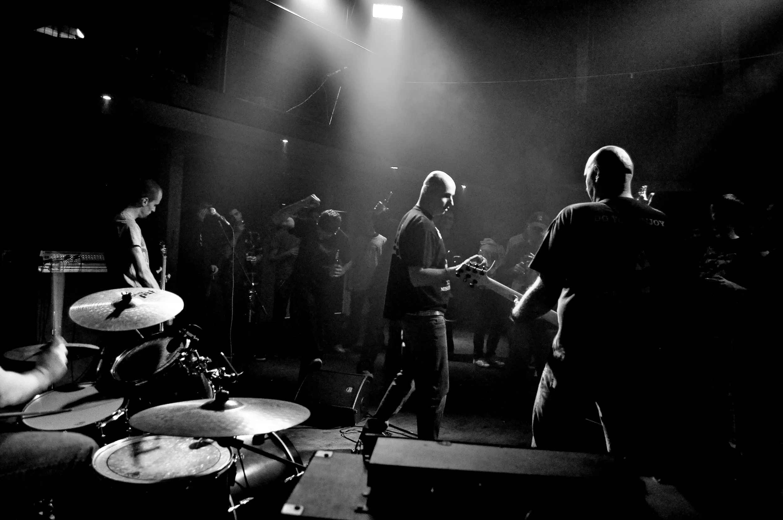 A group of people gathered in a dark indoor space, possibly a music venue or rehearsal room. There are musical instruments and microphones, and a person is playing drums in the foreground. The scene is illuminated by stage lighting creating a moody a