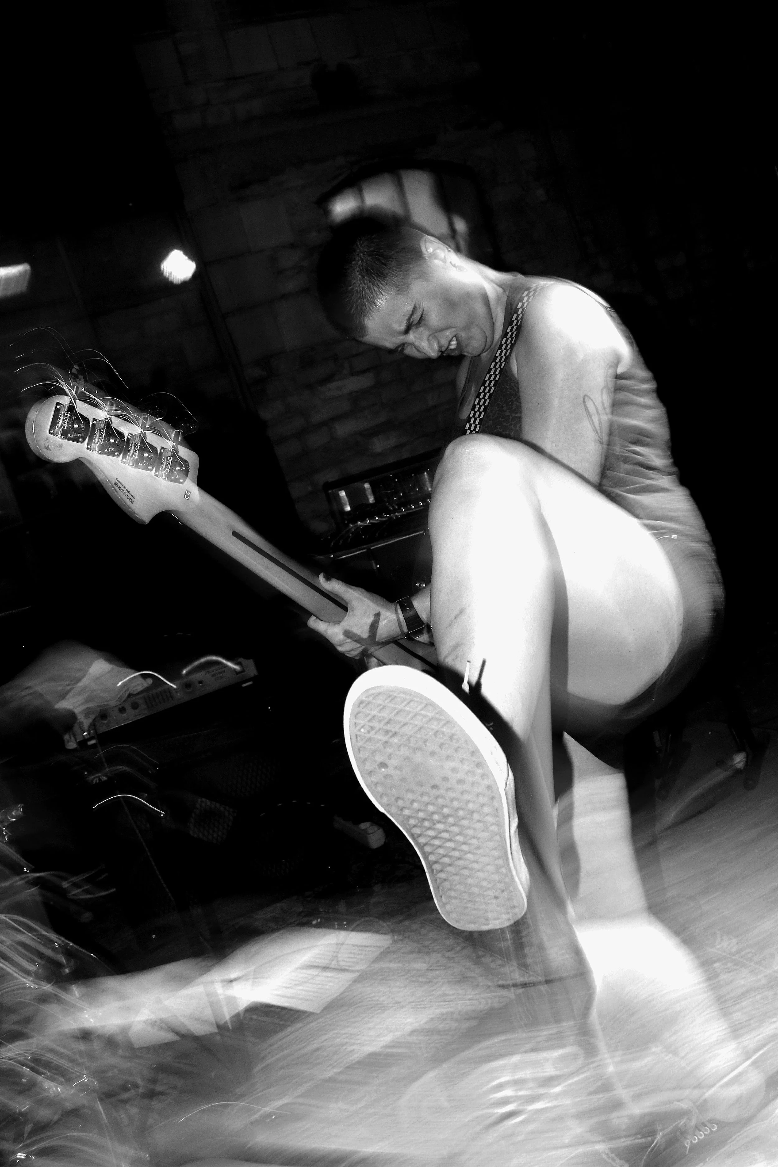 Black and white photo of a man with short hair playing an electric guitar, sitting on a chair in front of a brick wall, with a laptop and other equipment nearby.