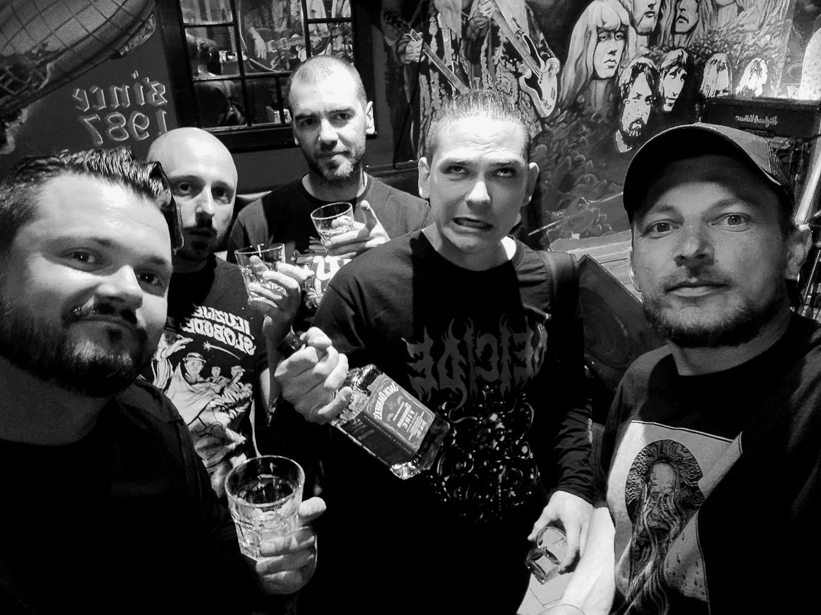 A black and white photo of five men in a bar or pub, holding drinks and posing for a group selfie. The background features a colorful mural with faces and musical instruments.