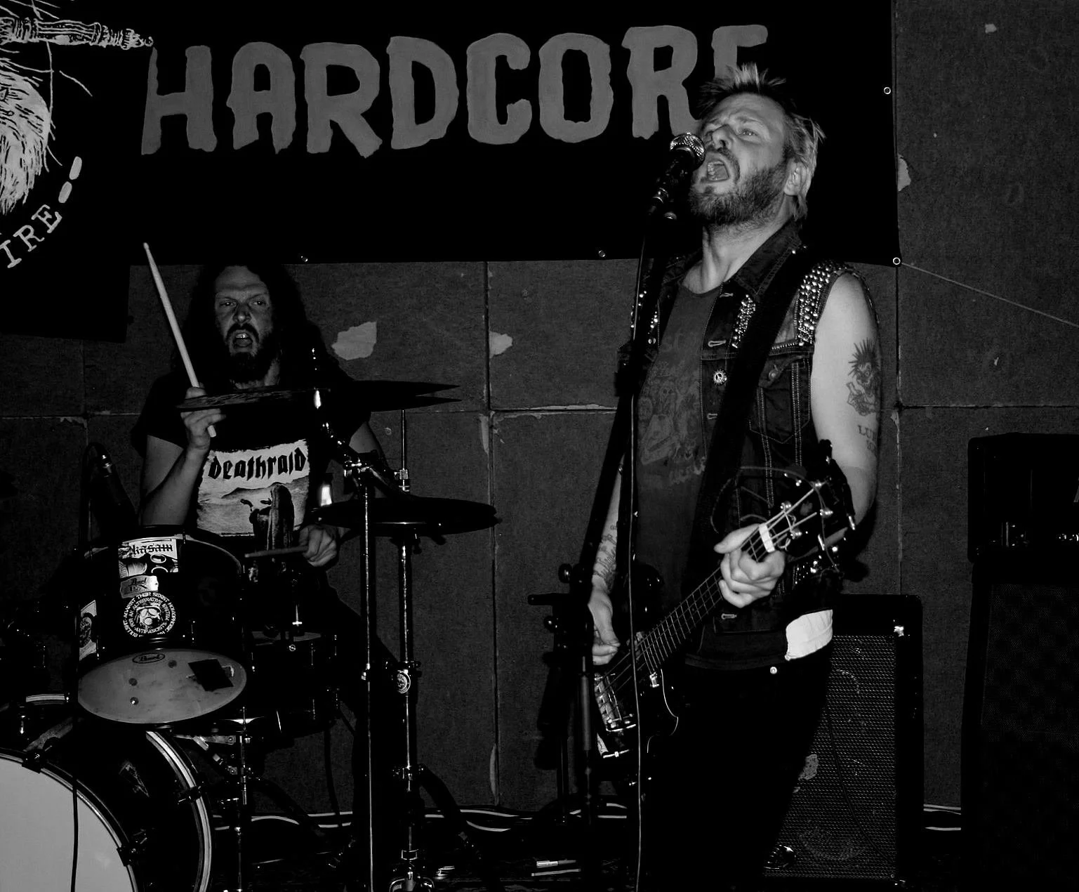 Two musicians performing on stage, one singing into a microphone and playing an electric guitar, the other playing drums, with a 'HARDCORE' banner in the background.
