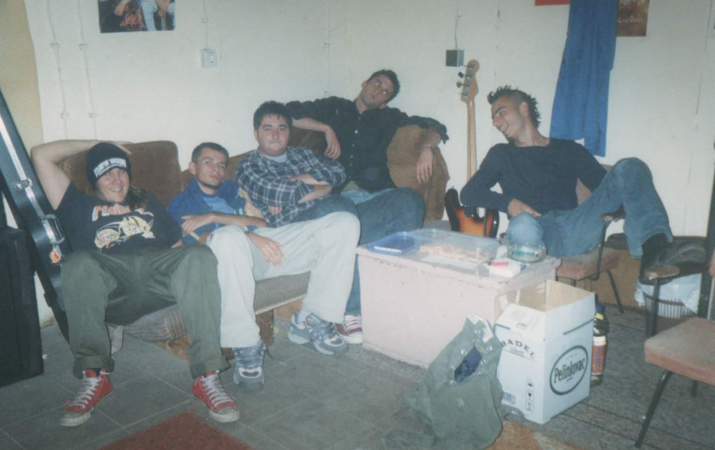 A group of five young men sitting in a room with beige walls, a brown couch, and a small table, appearing relaxed and casual. One is lying back with a beanie, three are seated on the couch with crossed arms or arms on their laps, and one is sitting o