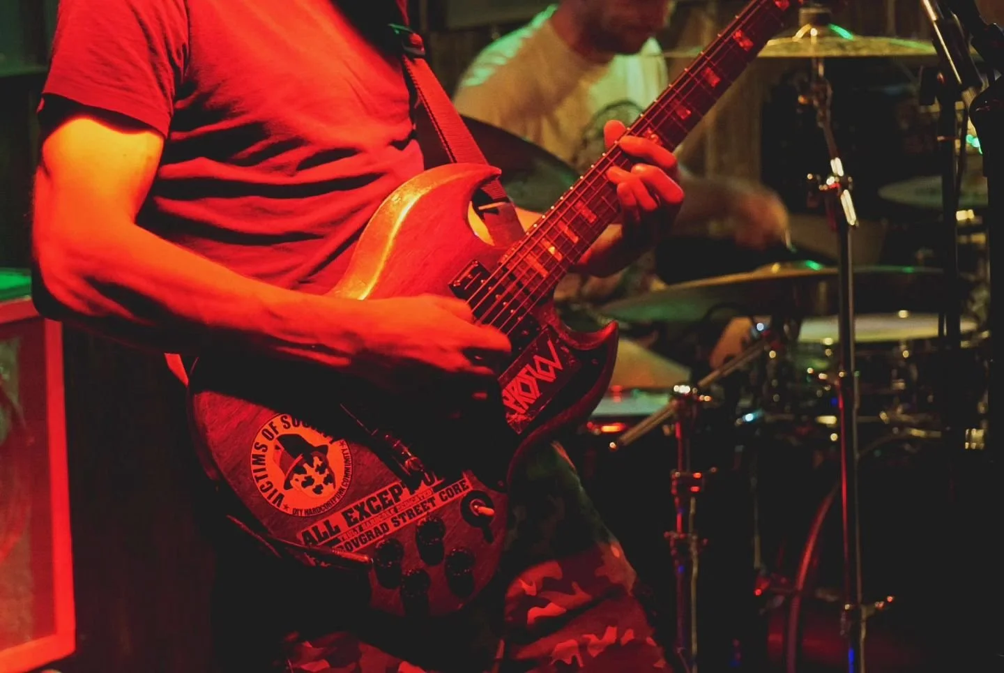 A person wearing a red shirt playing an electric guitar, with another person playing drums in the background, in a dimly lit setting with colorful lighting.
