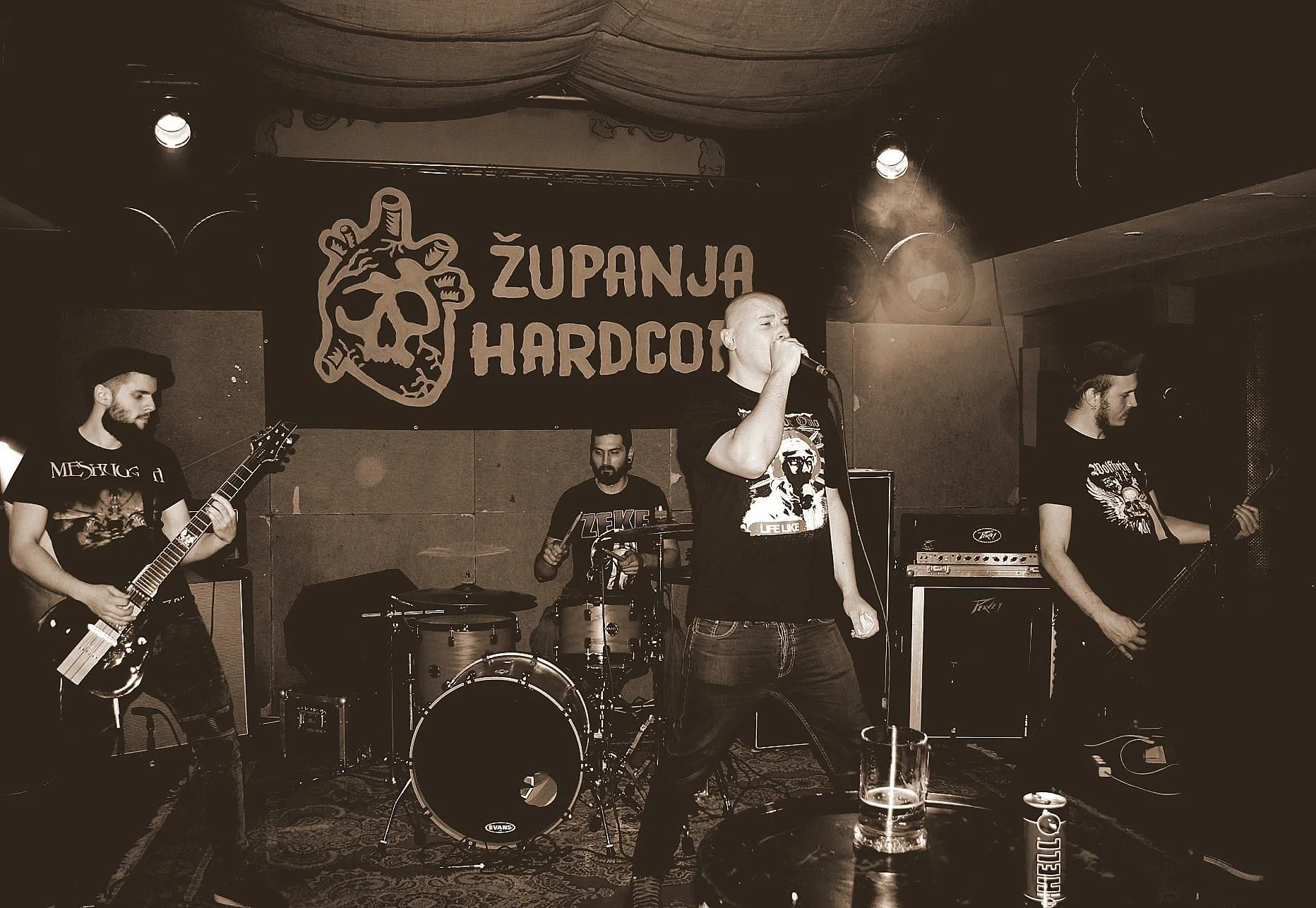 A band performing live on stage at a venue in front of Županja Hardcore flag, with four members: a guitarist on the left, a drummer behind, a vocalist in the center holding a microphone, and a bassist on the right. 
