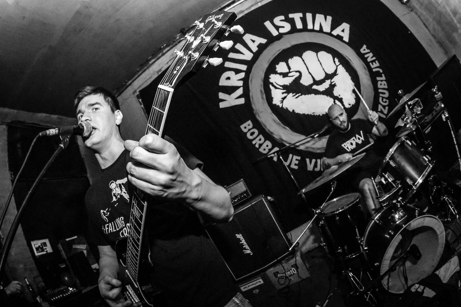 Black and white photo of two male musicians performing live on stage; one playing guitar and singing into a microphone, the other playing drums. Behind them, a circular sign reads "KRVIA ISTINA - BORBA JE VJEČNA"