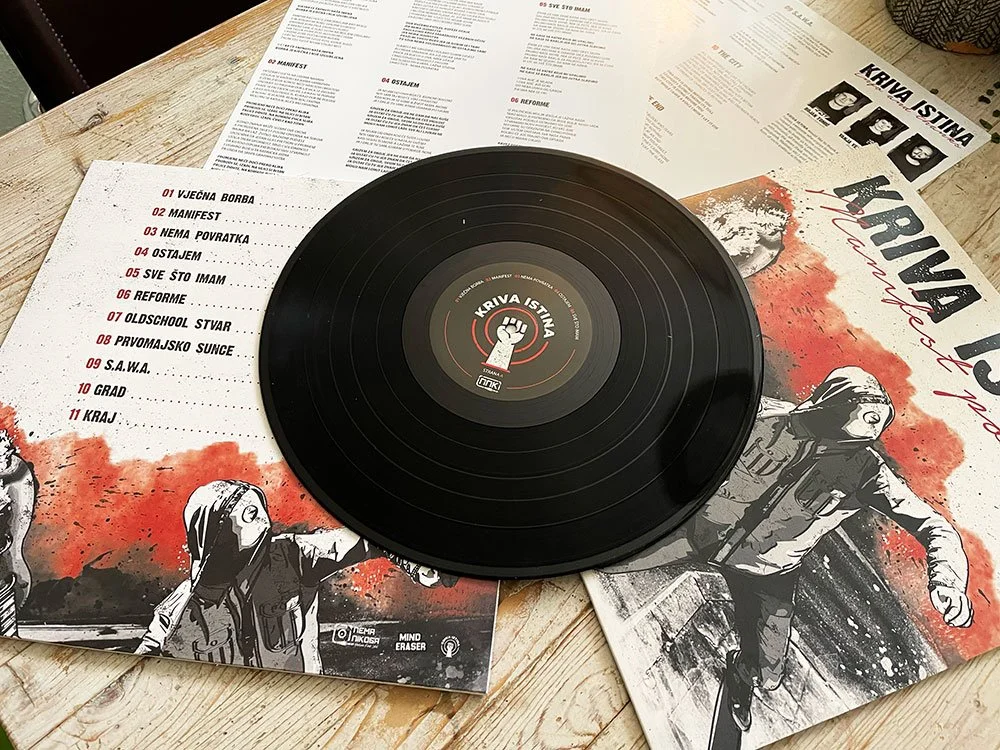A black vinyl record on top of printed pages and a magazine featuring artwork and text. The record label has a fist symbol and the name 'Kriva Istina'. The pages contain a tracklist in Serbian and show black-and-white illustrations of women wearing h
