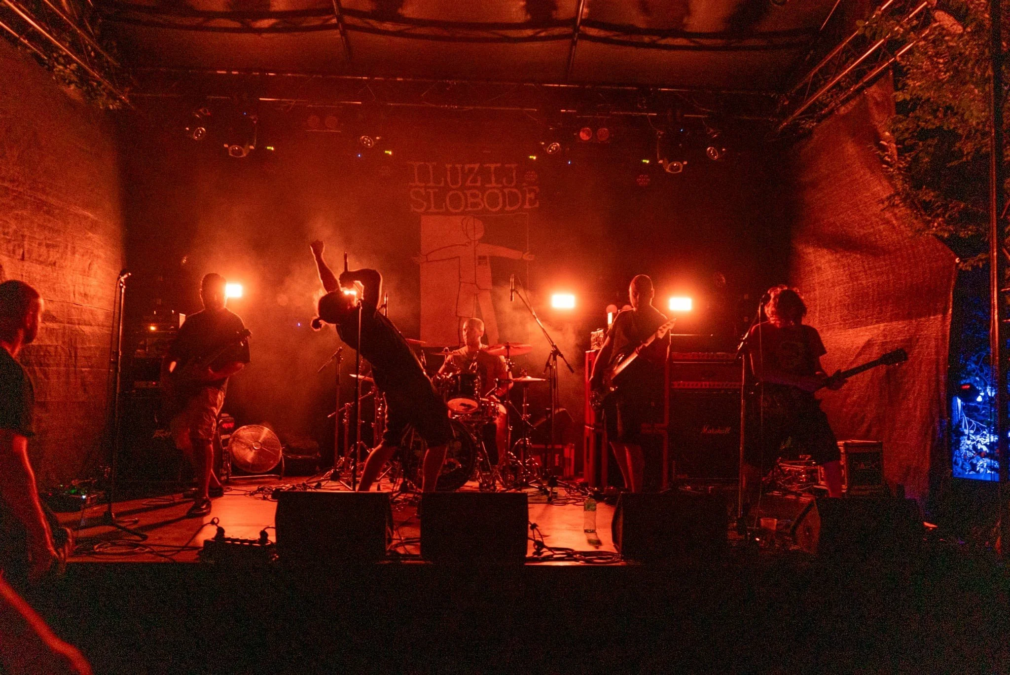 A band performing on stage at Luzid Slobode, with red stage lighting, musical instruments, and equipment, against a brick wall background.