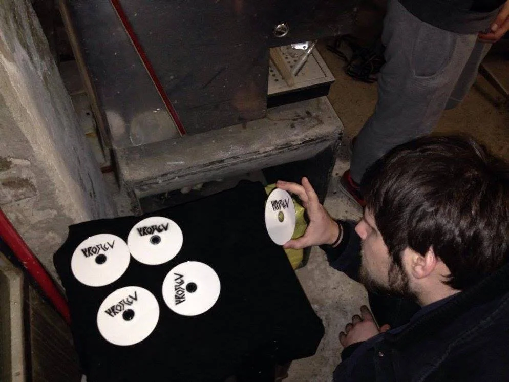 A man with brown hair and a beard is placing a CD with the label 'Proslov' into a black box, which already contains four other CDs labeled 'Proslov'. The setting appears to be a workshop or storage area with a workbench and other tools around.