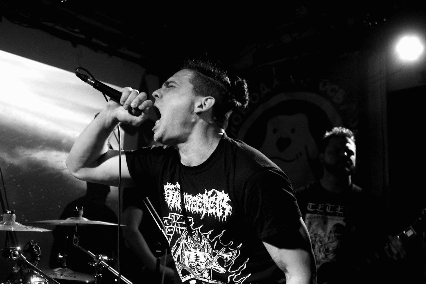Musician singing into a microphone during a live performance, with a drummer and guitarist in the background, in black and white.