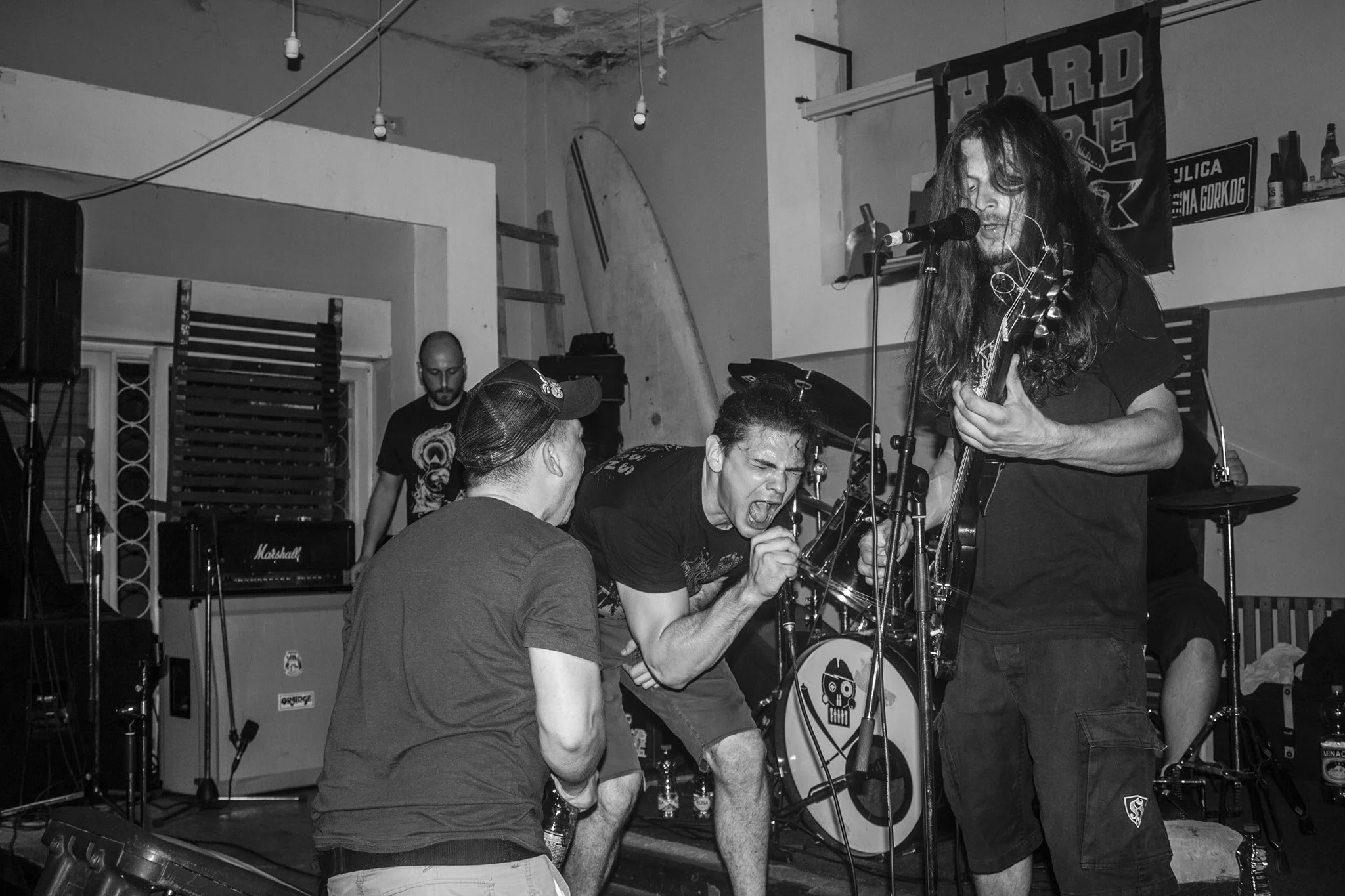 A black and white photo shows a band performing live. A guitarist is singing into a microphone while playing, and a man in a t-shirt and cap is recording or adjusting something nearby. The background has musical equipment and a banner on the wall.