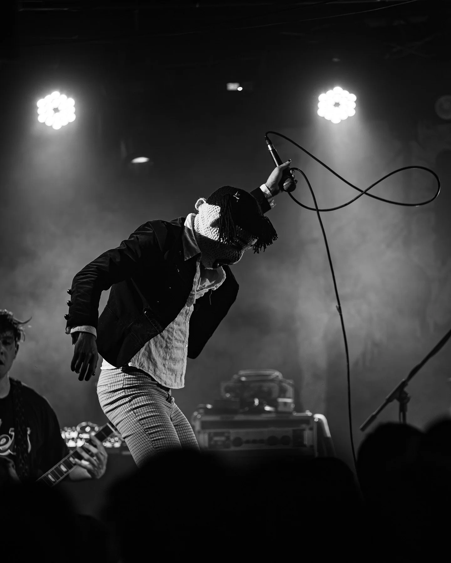 A performer on stage holding a microphone in one hand, wearing checkered pants and a jacket, with stage lights illuminating the background.