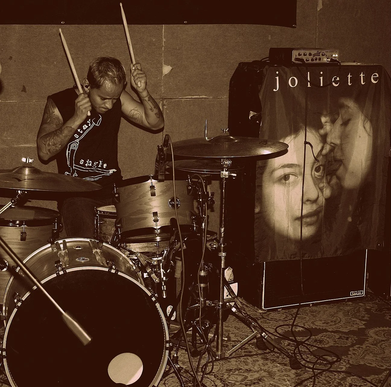 A drummer playing a drum set on stage with a large poster of a woman and child with the word 'Joliette' in the background.