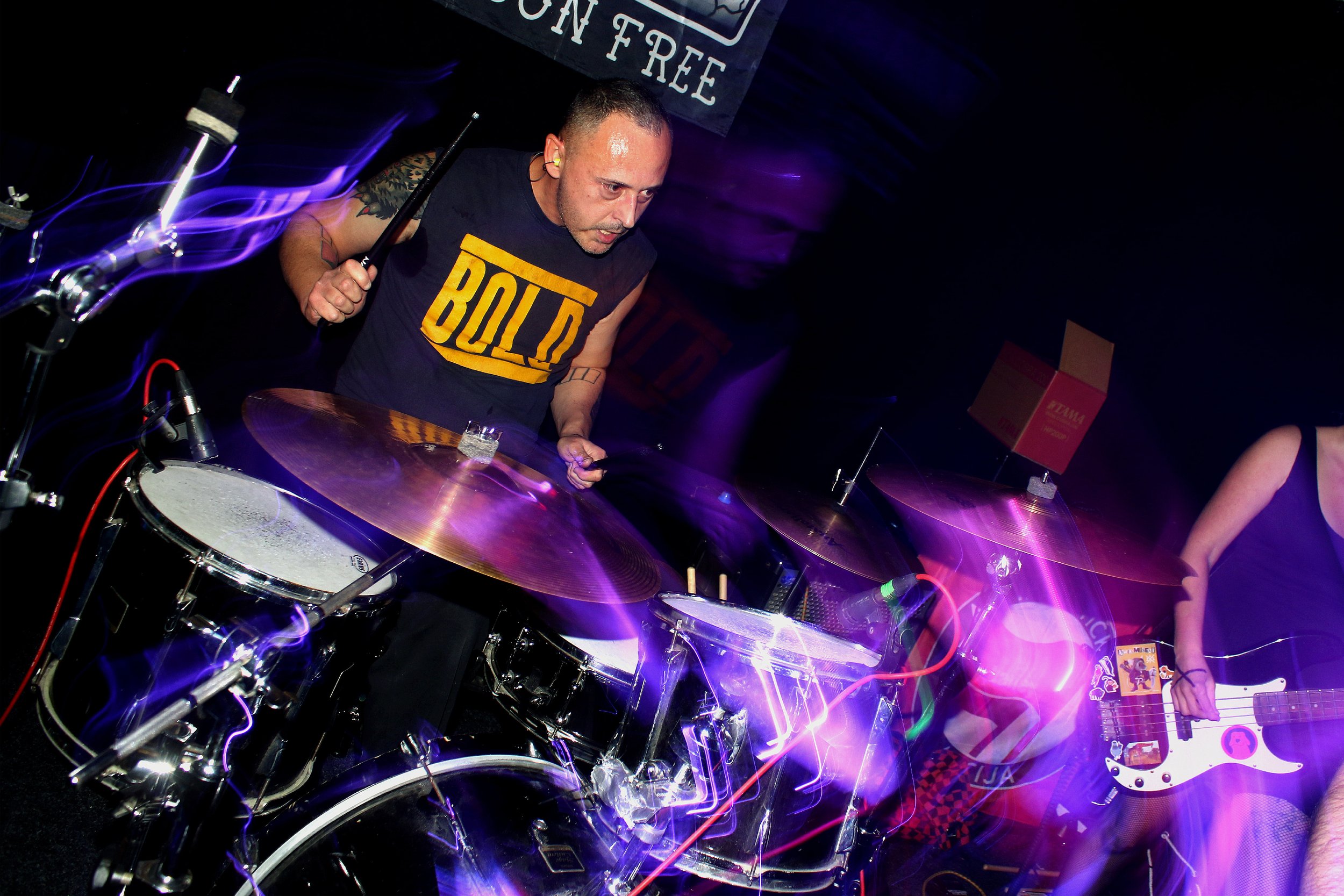 A man playing drums at a music event with purple light effects and a black background.
