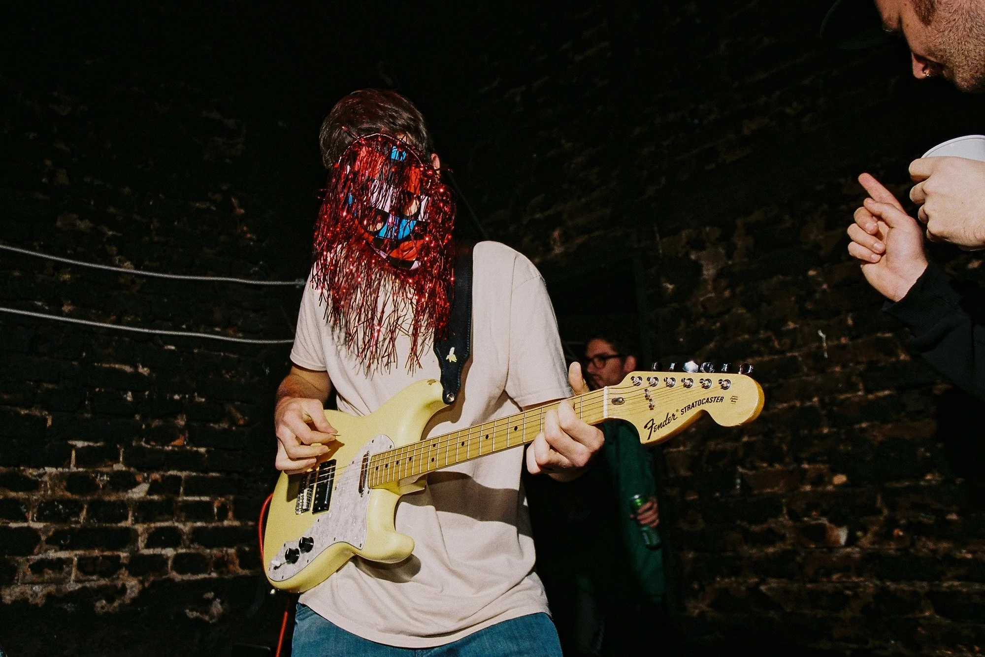 A person playing an electric guitar wearing a red tinsel mask, with a dark brick wall in the background.