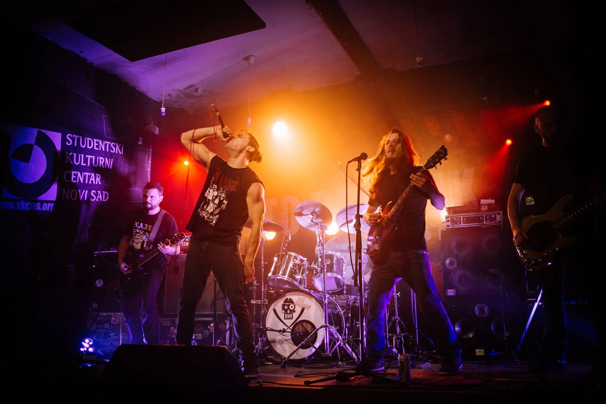 A live band performs on stage with a drummer, two guitarists, and a vocalist, under colorful stage lighting at an indoor music event.