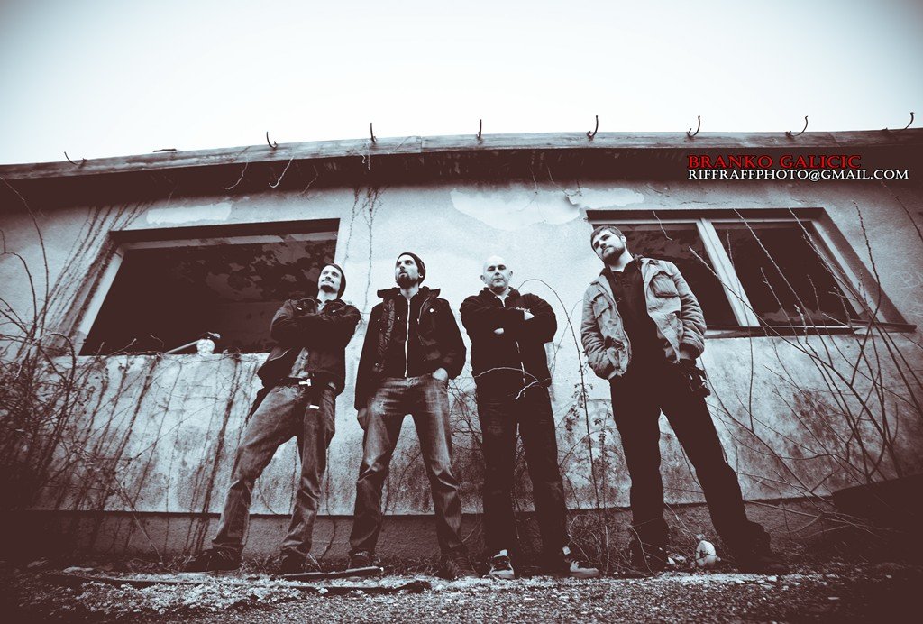Four men standing in front of an abandoned, dilapidated building with broken windows and around vine growth, looking confidently at the camera.
