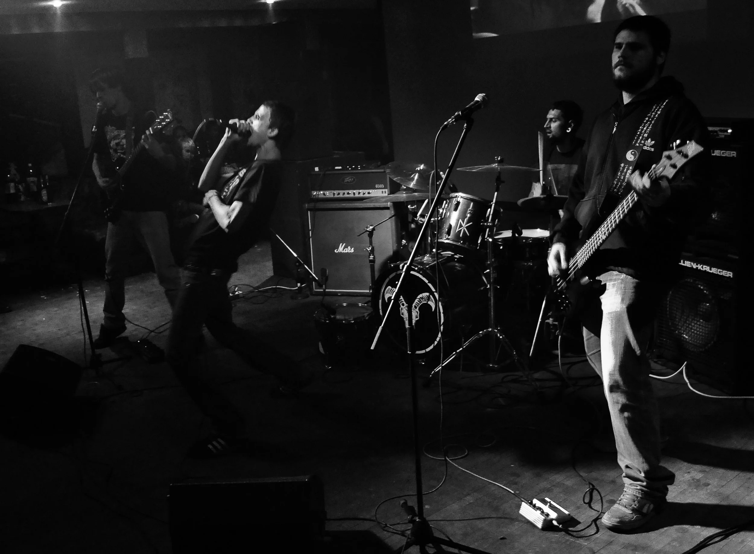 A band performing live on stage in a dark, intimate venue. The band includes a singer, guitarists, a drummer, and a bassist, with instruments and equipment around them.