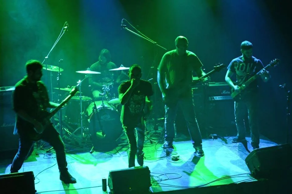 A five-member band performing on stage with green and blue lighting, including guitarists, a drummer, and a singer.
