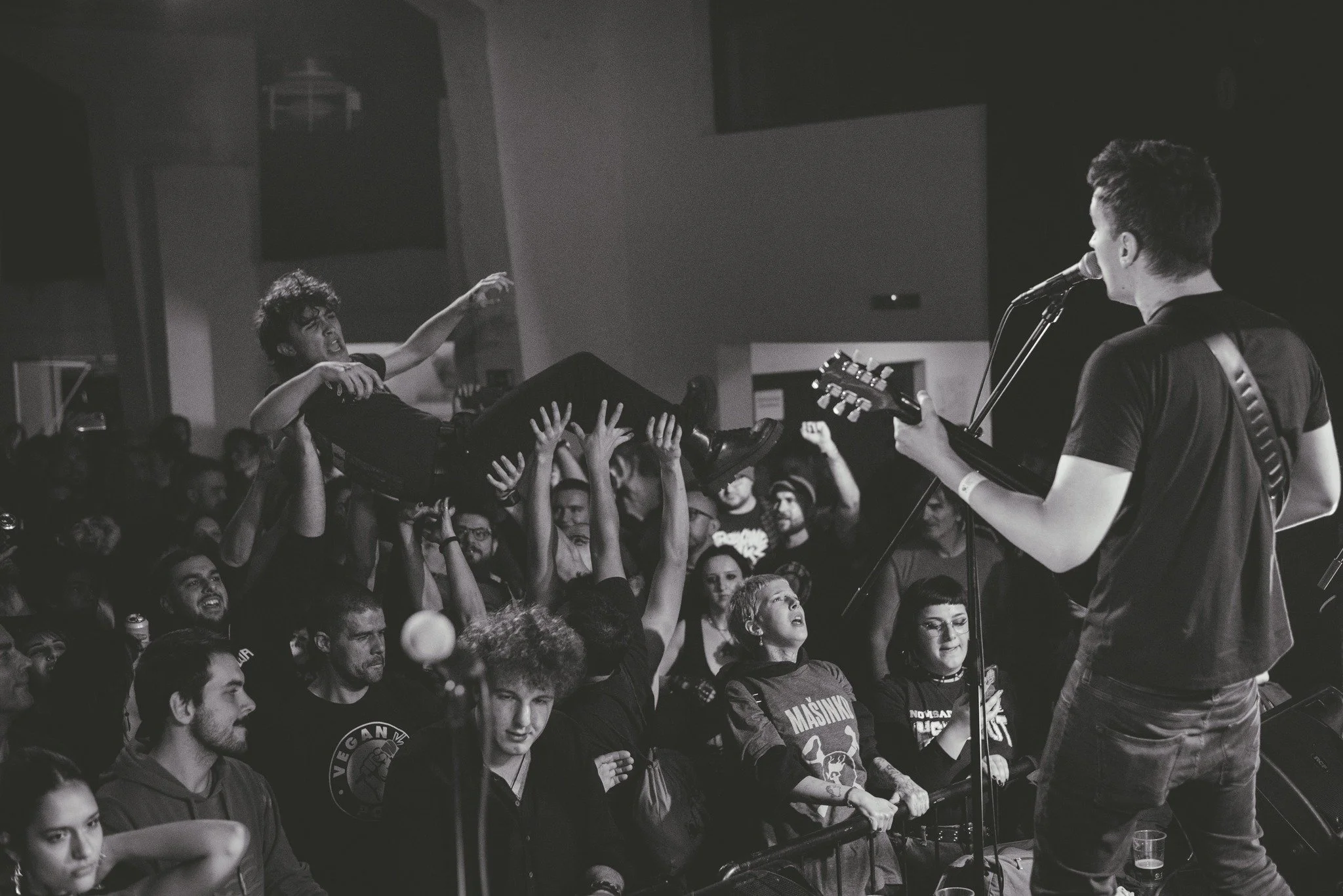 A black and white photo of a live music concert. A singer is holding a guitar and singing in front of a crowd, with band members nearby. Audience members are reaching out and looking at the performer.