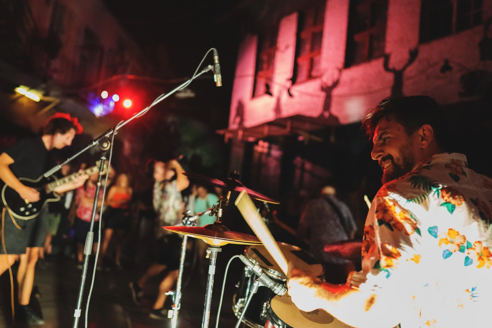 Musicians playing drums and guitar during a nighttime outdoor concert with an audience watching.