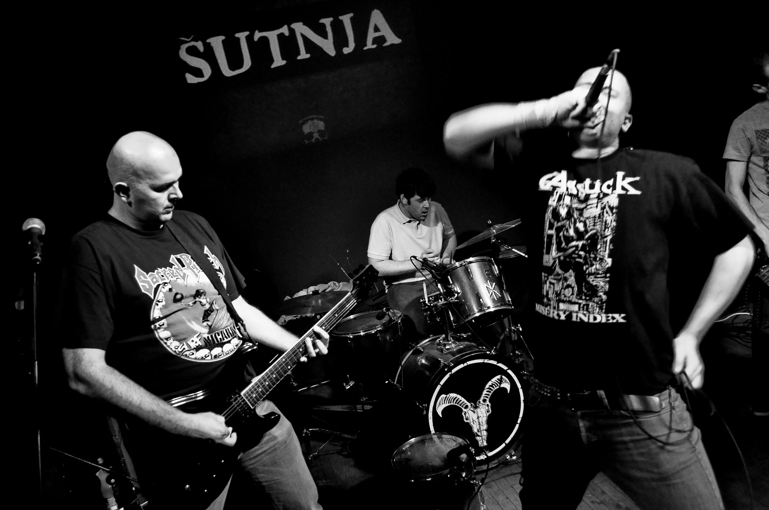 A band performing on stage, with a guitarist playing and a drummer behind, and the words 'SUTNJA' in the background.