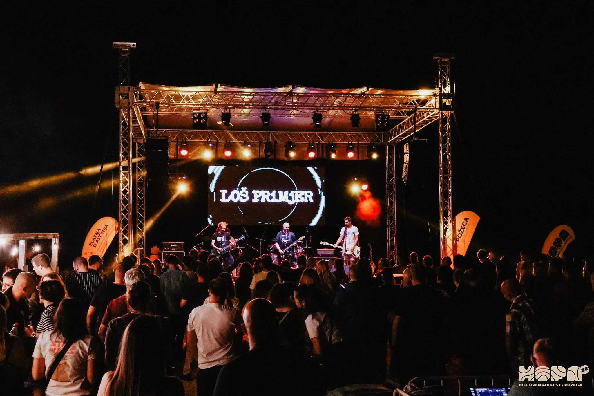 A live music performance on a stage at night with band members playing instruments and singing in front of a large crowd. The stage features a sign that reads 'LOS PRIMER' and lighting equipment.