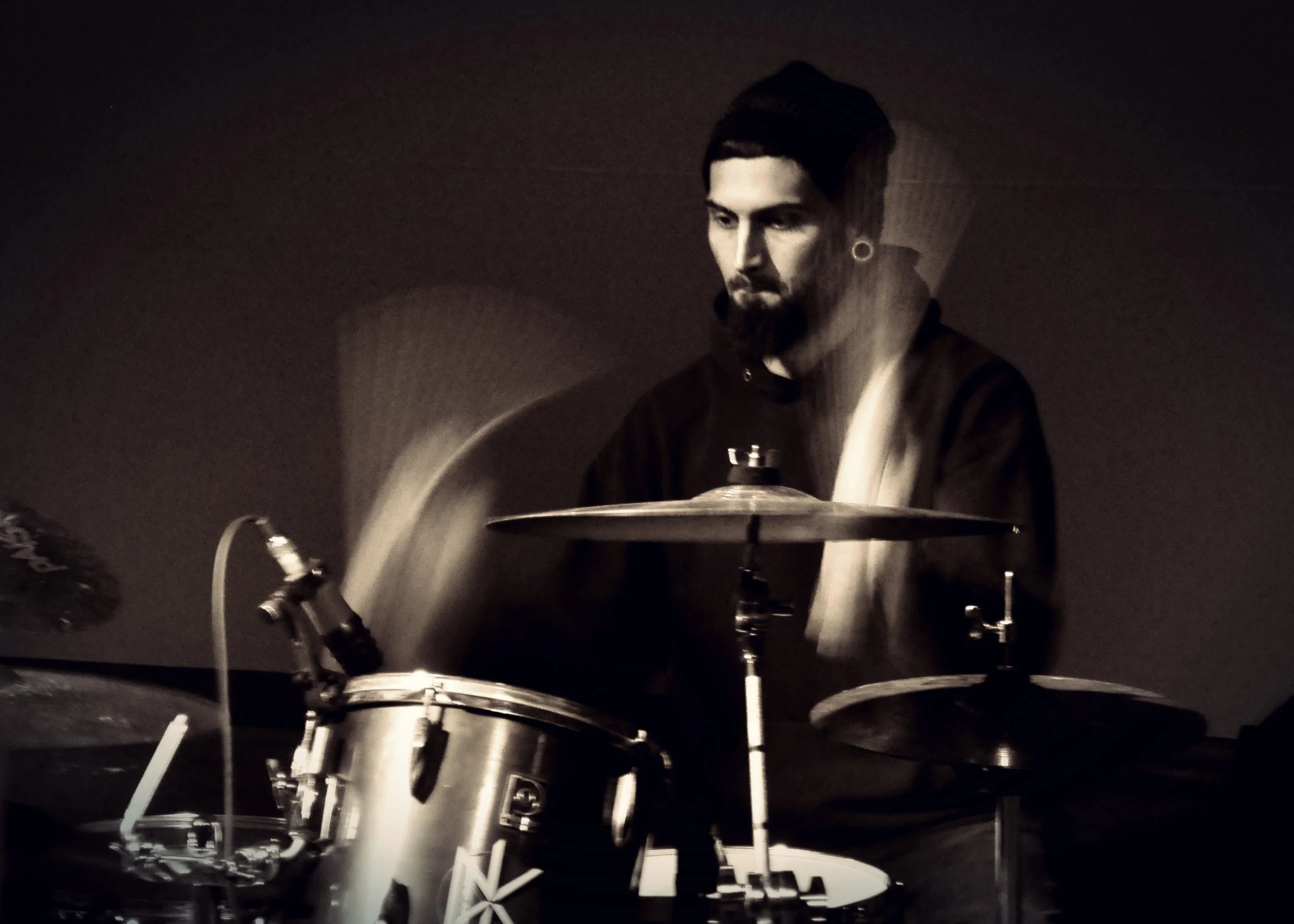 A person with dark hair and a beard, wearing a hoodie and a beanie, is playing his drum set in a dimly lit room.